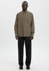 Brązowa koszula w kratę z rozchylanym kołnierzykiem, długimi rękawami i luźnym krojem, zestawiona z czarnymi spodniami i czarnymi butami.