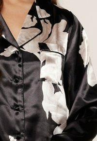 Chemise en satin noir avec motif floral blanc, poche poitrine unique et col cranté. Dotée de boutons sur le devant et de manches longues.