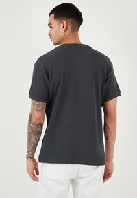 T-shirt grigio scuro con texture a waffle, colletto rotondo, maniche corte e vestibilità rilassata, indossato da una persona con tatuaggi sul braccio.