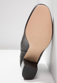 Černá kožená dámská bota na vysokém podpatku s texturovaným gumovým spodkem, čtvercovým podpatkem a ozdobnými stříbrnými nýty na straně.