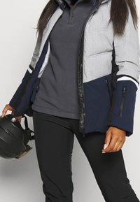 Isolerad jacka med ett grått och marinblått färgblock-design, dragkedjefickor och hög krage, i kombination med en grå sweatshirt och svarta byxor.