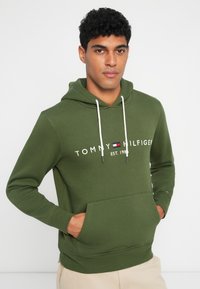 Zöld kapucnis pulóver, puha anyagból készült, elülső zsebbel, húzózsinórral ellátott kapucnival, és fehér logó felirattal kék és piros kiegészítőkkel.