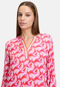 Bunte pinkfarbene Bluse mit geometrischem Muster, das rote und hellpinkfarbene Akzente aufweist, einen V-Ausschnitt mit kontrastierenden Streifen und langen Ärmeln.