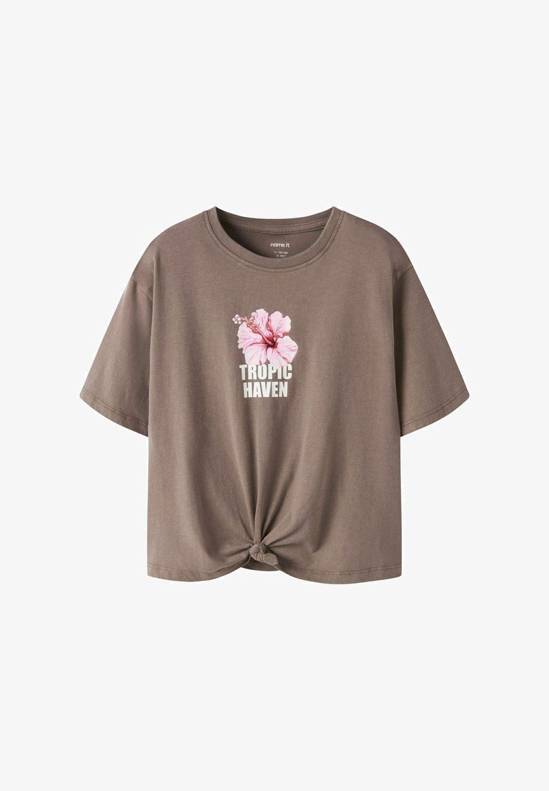 Καφέ κοντομάνικη μπλούζα με κόμπο μπροστά, γραφικό ροζ άνθος ιβίσκου και λευκό κείμενο "TROPIC HAVEN" στο κέντρο του στήθους.