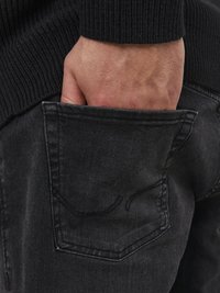 Vaqueros de mezclilla negra con un acabado texturizado, mostrando una mano en el bolsillo trasero. El bolsillo presenta detalles de costura discretos.