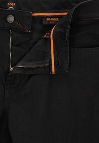 Jean slim noir avec coutures orange, fermeture éclair partiellement ouverte, bouton marqué "BOSS" et étiquette de marque "DELAWARE SLIM FIT" visible à l’intérieur de la ceinture.