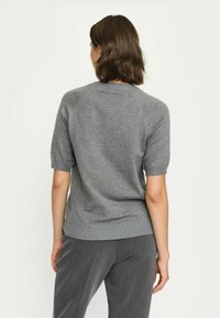 Vrouw die met haar rug naar de camera staat, draagt een grijze trui met korte mouwen en grijze broek tegen een effen lichte achtergrond.