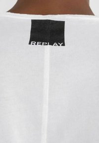 Maglietta bianca con etichetta nera con la scritta "REPLAY" sul retro. Il tessuto è liscio e il colletto è rotondo con cuciture visibili.