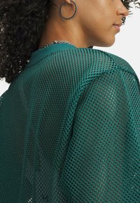 Zelena mrežasta majica ohlapnega kroja z grobo teksturiranim vzorcem mreže. Tkanina je polprosojna in lahka, z okroglim vratnim izrezom.