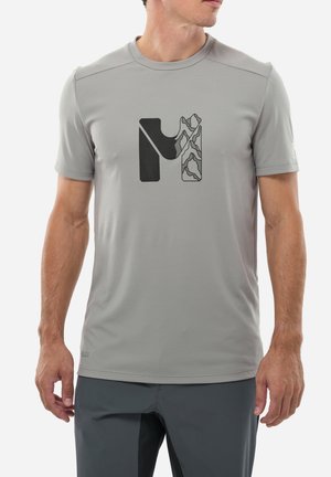 Homme portant un t-shirt gris clair avec un graphique abstrait de montagne en noir et gris sur la poitrine et un pantalon gris foncé.