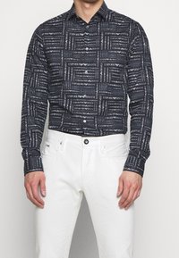 Camisa de manga larga en azul oscuro adornada con un patrón repetido de logotipos blancos; cierre frontal con botones y cuello clásico. Combinada con pantalones blancos.