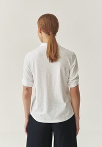 Biała koszula z guzikami i podwiniętymi rękawami, wykonana z lekkiego materiału. Posiada zaokrąglony dół i prosty kołnierzyk. Brak widocznych wzorów.