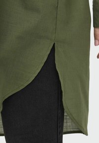 Vêtement long olive avec un ourlet arrondi et des fentes sur les côtés, fabriqué en tissu texturé, associé à des leggings noirs.