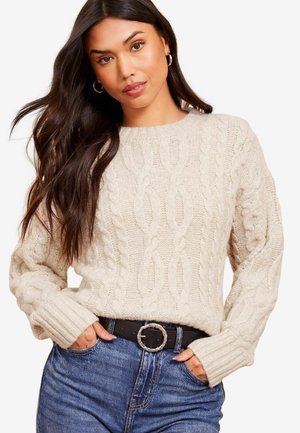 Helles beiges, grobstrickiges Sweatshirt mit rundem Ausschnitt und gerippten Manschetten, kombiniert mit hochgeschnittenen blauen Jeans und einem schwarzen Gürtel.