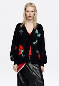 Czarny kardigan z kwiatową haftem w kolorach czerwonym, morskim i białym, z długimi rękawami i zapinany na guziki, noszony na czerwonej bluzce i czarnej spódnicy.