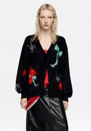 Cárdigan negro con bordado floral en rojo, verde azulado y blanco, con mangas largas y cierre de botones, combinado con una blusa roja y una falda negra.