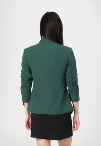Zelený přiléhavý blazer s řasenými rukávy, šitý na míru, s hladkou texturou. Nosený přes černou sukni, která vystihuje moderní, strukturovanou siluetu.