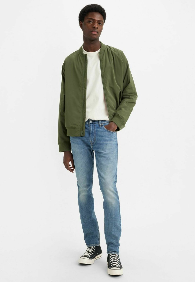 Veste bomber olive en tissu lisse, t-shirt blanc à col ras du cou, jean bleu clair et baskets noires avec semelles blanches.