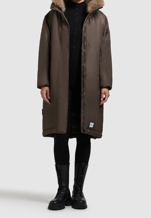 Personne portant un long manteau matelassé vert olive avec capuche bordée de fourrure, pantalon noir et bottes de combat noires à lacets, debout devant un fond uni.