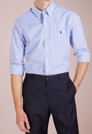 Homme portant une chemise à carreaux bleue et blanche à manches retroussées et un pantalon bleu marine foncé, main dans la poche.