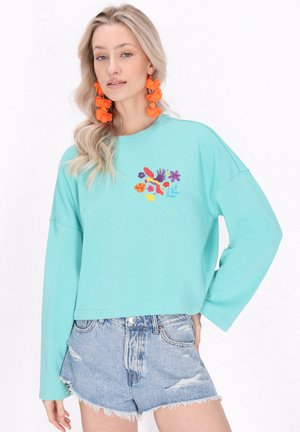 Turquoise langemouwen shirt met bloemenborduursel in rood, roze en blauw, gecombineerd met versleten denimshorts.