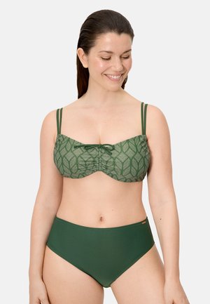 Mujer con un bañador de dos piezas verde con la parte superior estampada que tiene doble tirante en los hombros y braguitas altas de color sólido, sonriendo y mirando hacia abajo.