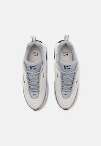 Tenisky Nike Air s krémovým vrchom, šedými akcentmi, textúrovanými prekrytiami a čiernou podrážkou. Obsahujú šnúrky a pútko na päte pre jednoduché obúvanie.