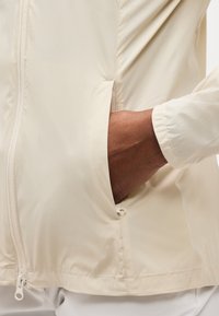 Ruka čiastočne vo vrecku svetlobéžovej prešívanej bundy na zips, s viditeľným ťaháčikom pri leme.