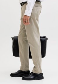 Mann iført beige bukser, svarte skinnsko, hvit skjorte, holder en svart skinnmappe mot en ensfarget bakgrunn.