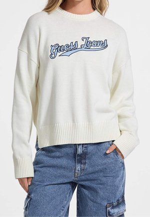 Kobieta nosząca biały sweter z dzianiny z niebieskim haftem "Guess Jeans", w połączeniu z niebieskimi jeansowymi spodniami typu cargo.