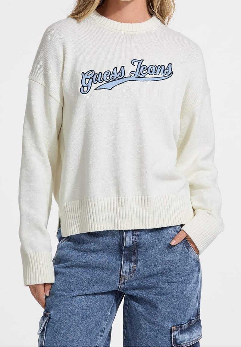 Femme portant un pull en maille blanc avec "Guess Jeans" brodé en bleu, associé à un pantalon cargo en denim bleu.
