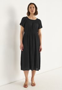 Schwarzes Midi-Kleid mit kurzen Puffärmeln, gerafftem Ausschnitt und taillierten Bund. Leichter, strukturierten Stoff fließt bis zum Knöchel.
