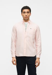 Homme aux cheveux courts et bouclés portant une chemise à boutons rose clair par-dessus un t-shirt rayé blanc et vert, ainsi qu'un pantalon foncé, debout devant un fond blanc.