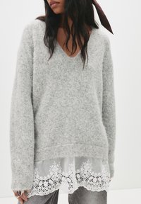 Szary dziergany sweter z dekoltem w serek, długimi rękawami i warstwową białą koronką na dole, ozdobiony wzorami kwiatowymi.