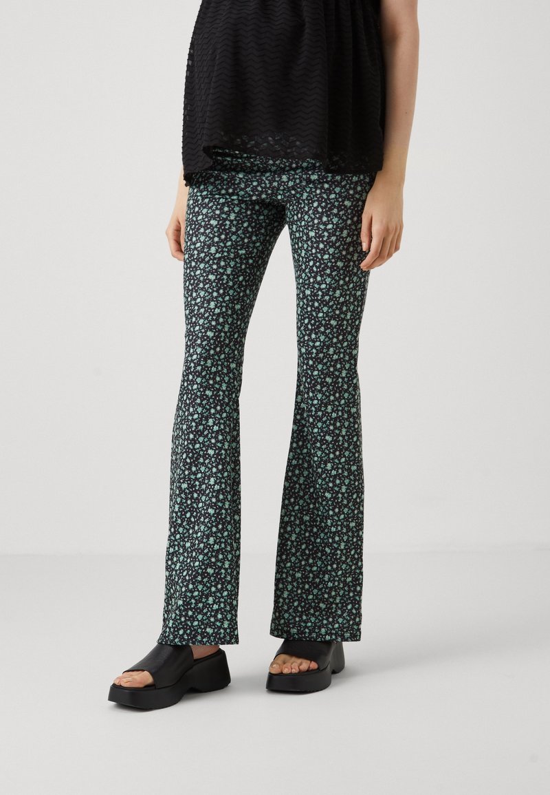 Pantalons évasés avec un fond noir et un petit motif floral vert, en tissu extensible. Portés avec des sandales noires épaisses.
