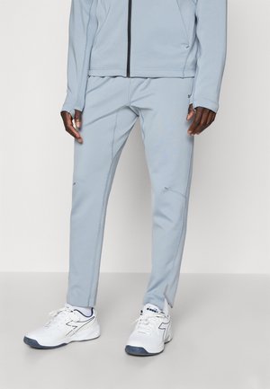 Pantalon de sport bleu clair avec aérations zippées aux chevilles, porté avec des baskets blanches et une veste à fermeture éclair assortie.