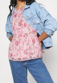 Růžový květinový top s peplum střihem, zdobený vzorem růží, spárovaný se světlemodrou džínovou bundou. Džíny jsou tmavě modré, což vytváří ležérní vzhled.