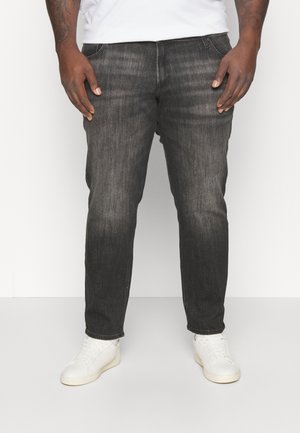 Hombre con vaqueros negros desgastados y zapatillas blancas, de pie frente a un fondo blanco liso con las manos relajadas a los lados.