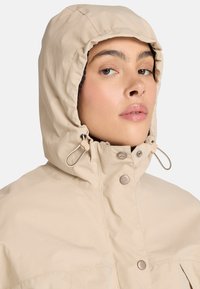 Chaqueta ligera beige que cuenta con una capucha ajustable con cordón, cierres a presión y un diseño minimalista con textura suave.