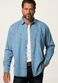 Camicia di jeans blu chiaro con maniche lunghe, due tasche sul petto e bottoni marroni. Indossata sopra una t-shirt bianca e jeans scuri.