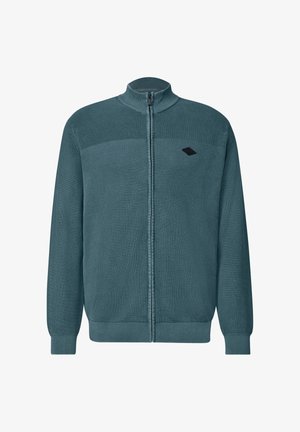 Homme portant un pull zippé côtelé bleu sarcelle par-dessus une chemise blanche, vu des épaules jusqu'au milieu du torse sur un fond uni.