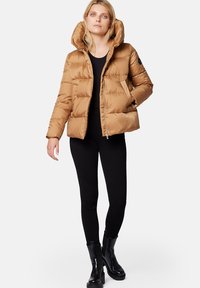 Kurtka puchowa w kolorze tan z odpinanym kapturem, pikowany design i zapięcie na napy, w połączeniu z czarnymi legginsami i błyszczącymi czarnymi botkami na kostkę.