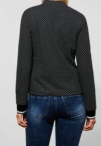Černá vzorovaná bunda s dlouhými rukávy a vysokým límcem. Má žebrované manžety s bílými pruhy, v kombinaci s modrými džíny.