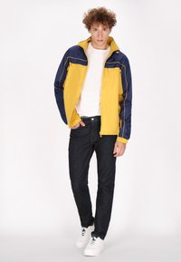 Geltonas ir tamsiai mėlynas vėjaraštis, su užtrauktuku, kontrastingais siūlais, derinamas su tamsiomis džinsomis ir baltais sportbačiais.