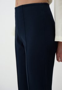 Pantalons taille haute bleu marine avec une texture lisse, présentant des coutures latérales soignées et un design ajusté, partiellement visibles à côté d'un haut blanc.