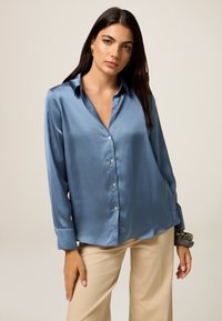 Blouse satin bleu clair avec un col, des manches longues et une fermeture à boutons. Texture lisse avec une coupe décontractée et des poignets retroussés.