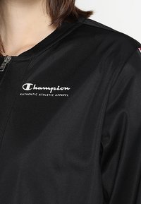 Svart sportjacka med dragkedja fram, med en "Champion"-logotyp och "Authentic Athletic Apparel" i vit text på bröstet. Slät textur.