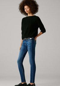 Mustast kudumist kampsun, sinised kitsad teksad ja mustad jalanõud. Märkimisväärsed detailid hõlmavad tekstuuri kampsunit ja naeltega rihma. Neutraalne taust.