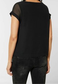 Svart topp med korta, uppvikta ärmar, i mjukt material med genomskinliga inslag. Bärs med mörka, strukturerade jeans som visar detaljer på baksidan.