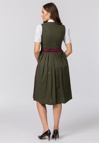 Grünes gemustertes Kleid mit einem taillierten Oberteil und einem plissierten Rock; verfügt über eine kastanienbraune Schärpe an der Taille; kombiniert mit schwarzen High Heels.